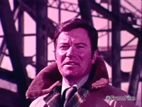 The still features a man, likely a prominent figure, standing in front of a large structure, possibly an industrial or urban setting. He has a serious expression and is dressed in a coat with a fur-lined collar, suggesting a cold environment. The colors appear vivid, with a slight pinkish hue dominating the scene. The context relates to a Public Service Announcement about the National Flood Insurance Program, highlighting the importance of protection against flooding for Americans.