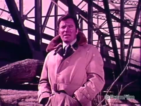 The still features a man, dressed in a tan coat with a fur collar, standing beneath a large bridge structure. He appears to be delivering an important message about the impact of floods on communities across America. The overall color tone has a vintage hue, indicative of the 1970s, setting a serious yet informative atmosphere. The context of the Public Service Announcement emphasizes the introduction of the National Flood Insurance Program as a means for people to protect themselves from flood-related damages.