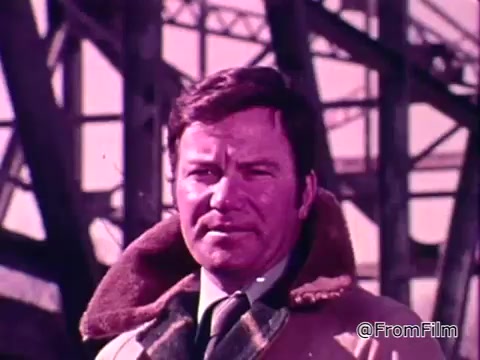 The still features a serious-looking William Shatner, dressed in a tan coat with a fur collar, standing in front of a large industrial structure. His expression conveys urgency and concern, likely reflecting the importance of the message regarding flood safety. The color palette has a vintage look, emphasizing the PSA's 1975 context. The overall tone suggests a call to action for viewers to consider the National Flood Insurance Program as a means of protection against flooding.
