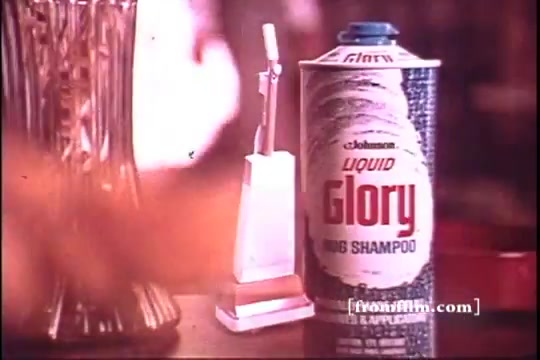 The still captures a vintage 1970s commercial for 'Liquid Glory Rug Shampoo.' Prominently featured is a spray can of the product, labeled 'Liquid Glory' alongside a white spray nozzle on a small stand. The background includes blurred elements typical of the era's aesthetic, giving a nostalgic feel to the scene. The color palette is warm, reflecting the style of advertisements from that time.
