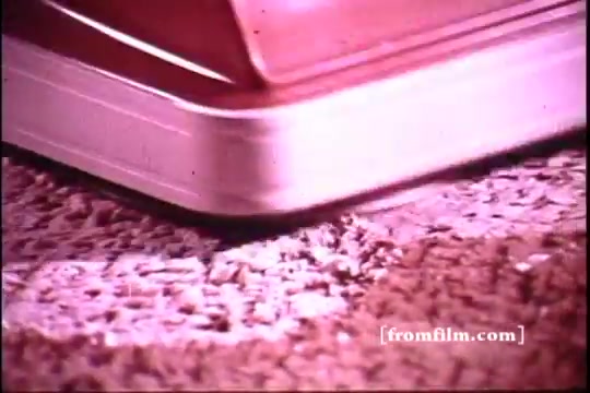 The still features a close-up of a rug cleaning device in action, focusing on its edge just above a textured carpet. The pinkish hue and graininess suggest it's from a 16mm film print, characteristic of 1970s commercials. The image captures a moment of cleaning, highlighting the product's use on carpeting.