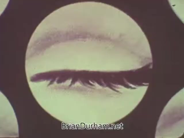 The still from the 'American Optometric Association PSA - 1960s' features a close-up view of an eye, focusing on the eyelid and eyelashes. The image is framed within a circular design, emphasizing the eye's contours. This visual likely aims to highlight the importance of eye care and vision health. The aesthetic is typical of the era, combining informative content with a visually striking presentation.