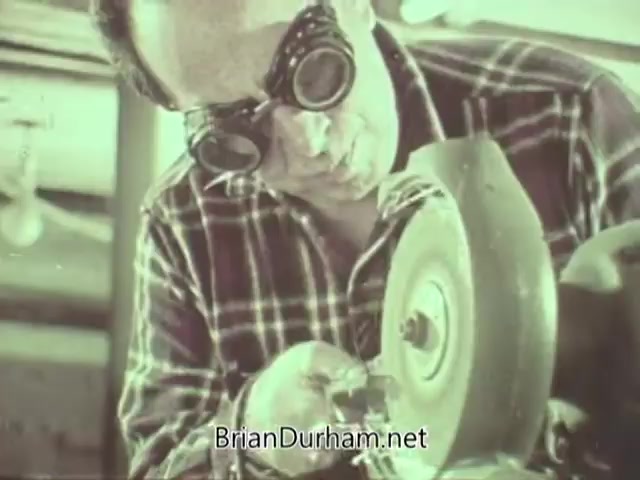 The still features a man wearing protective goggles, working intently on a machinery piece, likely related to eyewear or optical equipment. He is dressed in a plaid shirt and is using his hands to manipulate an object near a rotating tool. The image captures a moment focused on craftsmanship, highlighting the importance of safety in optometry-related work. The muted colors suggest a vintage aesthetic typical of the 1960s.