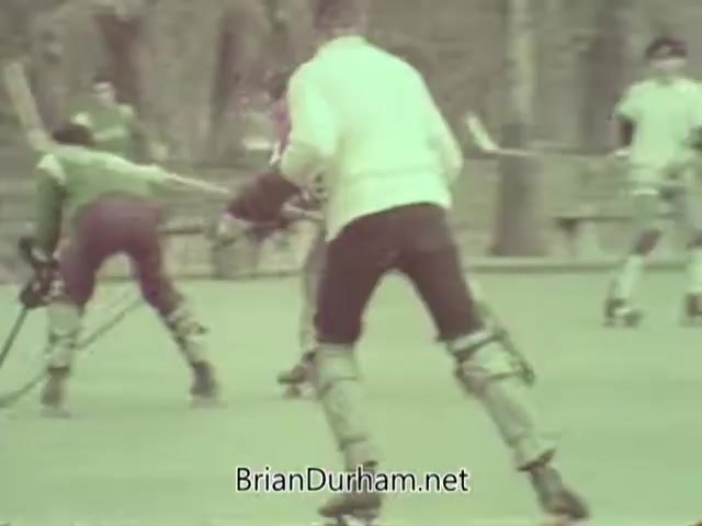 The still shows a group of players engaged in a roller hockey game, featuring individuals wearing protective gear and playing on a smooth surface. The players are dressed in various colored uniforms, with one notably in a white sweater. The background suggests an outdoor setting, likely a park or recreational area. The slightly muted colors evoke a vintage aesthetic typical of the 1960s.
