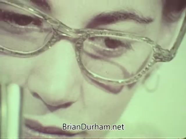 The still from the 'American Optometric Association PSA - 1960s' features a close-up of a woman's face, highlighting her glasses and attentive expression. She appears to be focused on an optometric task, possibly examining eyeglass prescriptions or conducting an eye examination. The image is indicative of the era, showcasing the emphasis on vision care and eye health awareness.