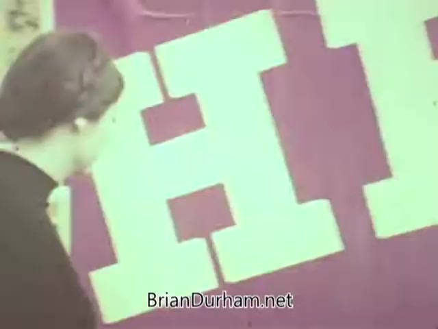 The still features a woman walking past a large, brightly colored banner with the word 'EYES' prominently displayed. The setting appears to be a public awareness event or campaign related to eye care, likely from the 1960s, reflecting the themes of health and optometry emphasized by the American Optometric Association. The woman's attire is typical of the era, hinting at the period's fashion.