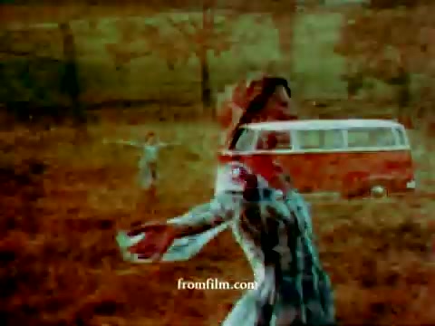 The 16mm still from the 1972 Volkswagen 'station wagon' commercial features a warm, vintage aesthetic with a couple in an outdoor setting. The woman is joyfully dancing, her arms open wide, while a red Volkswagen bus is partially superimposed in the background. Soft, muted colors create a nostalgic atmosphere, emphasizing carefree movements and a connection to nature. A child can be seen in the distance, contributing to the scene's playful vibe.