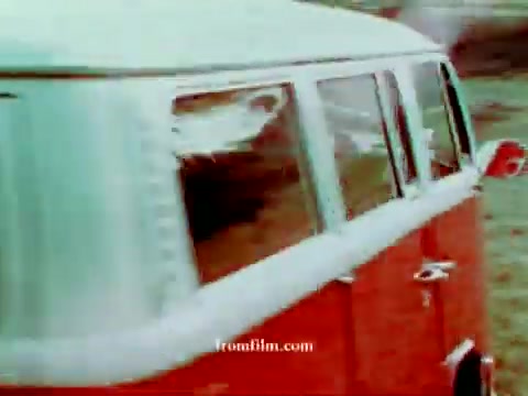 The still features a vintage Volkswagen station wagon, showcasing its orange and white exterior. The scene captures the vehicle from a side angle, emphasizing its iconic shape and design. The background appears blurred, suggesting motion or speed, while the color correction adds vibrancy to the image, enhancing its retro charm.