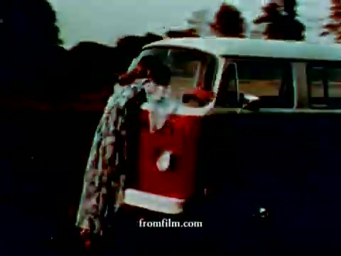 The still shows a vintage Volkswagen station wagon in a scenic outdoor setting. A figure is leaning against the vehicle, draped in a patterned blanket. The color correction has enhanced the vibrant hues, giving a retro feel to the image, typical of 1970s commercials. The overall mood is nostalgic, evoking a carefree, adventurous spirit associated with road trips.