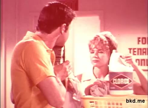 The still features a scene from a 1970s Clorox TV commercial, showcasing a man and a woman in a domestic setting. The woman, likely playing a homemaker role, is holding a bottle of Clorox and looking at the man, who appears engaged in conversation. The color palette is warm, typical of the era, with a slight pinkish tint. A basket of laundry is positioned in the foreground, further emphasizing the cleaning theme of the advertisement.