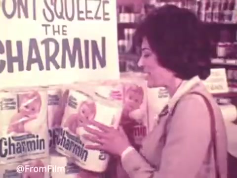 In this 16mm still from a 1970s Charmin commercial, a woman is seen smiling and engaging with a display of Charmin toilet paper. The prominent sign reads 'DON'T SQUEEZE THE CHARMIN,' emphasizing the playful nature of the ad. The woman appears enthusiastic, and the packaging features a baby, adding a touch of charm to the product's branding. The scene captures the light-hearted and friendly tone characteristic of advertising from that era.   .