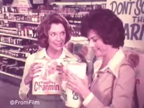 The still from the 'Charmin Commercial - 1970s' features two women in a grocery store, each holding packages of Charmin toilet paper. One woman, with curly hair, appears to be enthusiastically sharing information, while the other, dressed in a more formal outfit, looks engaged and interested. The background displays grocery shelves filled with products, setting a lively market scene. A sign can be seen that says 'DON'T SQUEEZE THE CHARMIN.' The image has a vintage quality typical of 16mm film from that era.