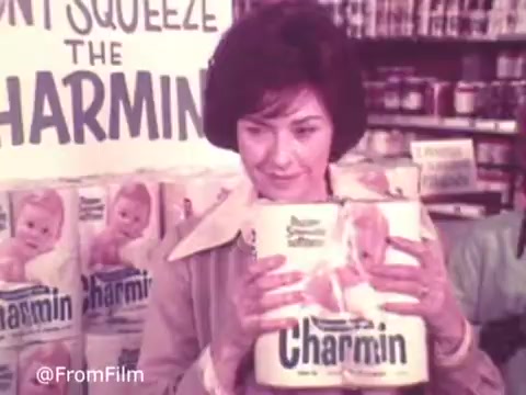 The still from the 1970s Charmin commercial features a woman smiling as she holds packages of Charmin toilet paper. Behind her, a prominent display reads 'Don't Squeeze the Charmin,' emphasizing the product's appeal. The warm tones and nostalgic style of the 16mm print capture the era’s advertising charm, showcasing the playful approach to marketing household products.