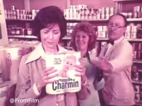 This still from a 1970s Charmin commercial features three characters in a grocery store aisle. The central figure, a woman, is examining a roll of Charmin toilet paper, while two others, a woman and a man, engage with her. The setting is cluttered with various products, conveying a typical supermarket environment of the era. The color palette and clothing reflect the fashion trends of the time. The commercial likely aims to highlight the product's qualities in a humorous or engaging way.