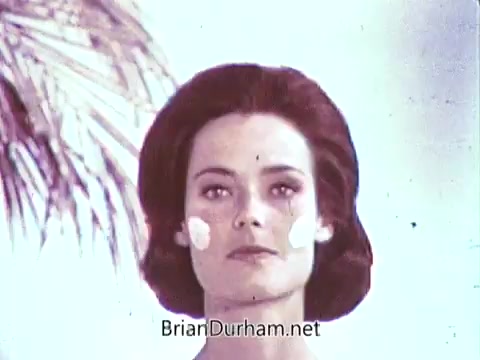 The still from the 1968 Dove Soap commercial features a woman with a smooth complexion, wearing natural makeup and a classic hairstyle. She is looking directly at the camera, with small dollops of cream or soap on her cheeks, suggesting a focus on skin care and beauty. The background is bright and airy, contributing to a fresh, clean aesthetic that aligns with the Dove brand messaging of gentleness and care.