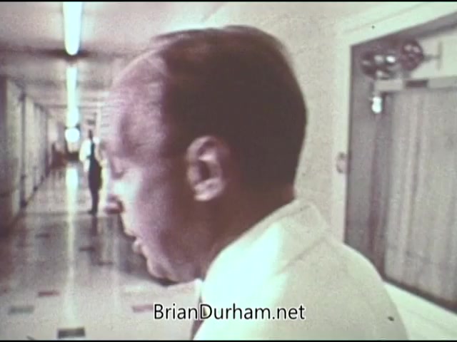 The still from the 1969 American Cancer Society PSA features a man in a white coat, likely a medical professional, walking through a hospital corridor. The setting appears clinical, with doors on either side and tiled flooring. The focus seems to be on the individual, who appears to be engaged in thought or conversation, contributing to the educational message about cancer awareness.