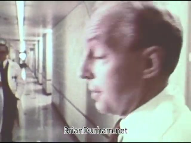 The still shows two men in a hospital setting. One man, in the foreground, appears to be wearing a white lab coat and has a serious expression. The background features a corridor typical of a medical facility, with lights overhead and more individuals in white coats visible, suggesting an environment focused on healthcare and potentially addressing cancer prevention or treatment. The overall tone reflects the clinical and educational nature of a public service announcement from 1969.