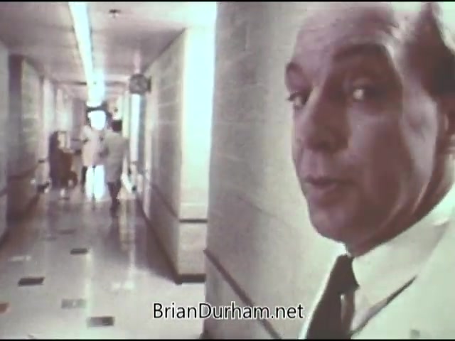 The still features a man in a suit, looking back toward the camera in a hospital corridor. He appears to be speaking or reflecting on a serious topic. In the background, medical professionals are seen walking, emphasizing the setting's clinical atmosphere. The image likely captures a moment intended to raise awareness about cancer, consistent with the American Cancer Society's messaging from 1969. The lighting and decor suggest a dated yet formal healthcare environment.