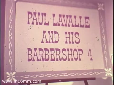 The still features a sign that reads 'PAUL LAVALLE AND HIS BARBERSHOP 4,' displayed in bold, purple lettering. The sign is framed with decorative elements, suggesting a festive or promotional context. This image likely highlights a performance or event related to Paul Lavalle and his group, fitting within the theme of community engagement promoted by The Boys Club PSA from 1968. The color and styling reflect the aesthetic of the time period.
