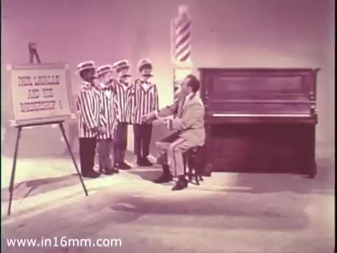 The still from the 1968 PSA for The Boys Club features four boys in striped jackets standing in front of a seated man, who is likely delivering a message or lesson. The scene includes a large sign with text, possibly outlining the club's mission or activities, and a piano nearby, suggesting a focus on music or performance. The overall tone appears educational and inviting, aimed at encouraging participation in the Boys Club.