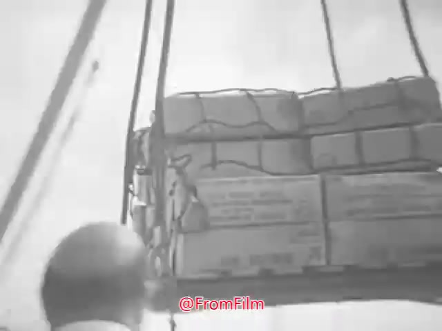 The still from the 16mm print of 'Project CARE PSA - 1960s' shows a man, likely a dock worker or overseer, looking at a large crate being lifted by a crane. The crate is secured with netting and appears to contain supplies, possibly aid-related goods. The setting suggests a port or loading dock, characterized by an industrial atmosphere. The image is in black and white, reflecting the film's era.