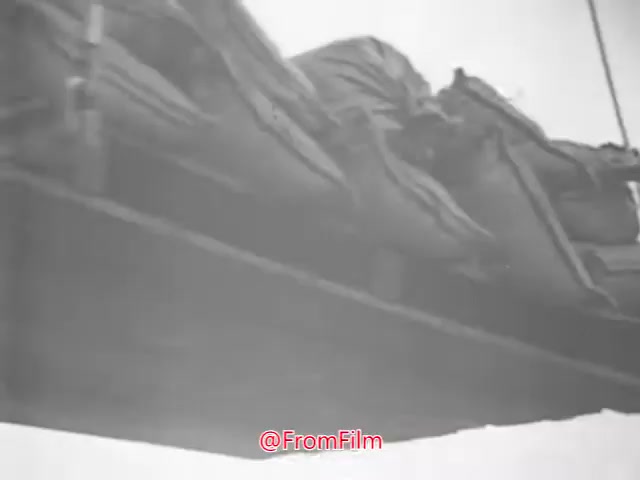 The still from the 16mm print of 'Project CARE PSA - 1960s' depicts a cargo delivery scenario, likely involving the transportation of supplies. The image shows a perspective of large, wrapped parcels suspended from a structure, hinting at an aerial drop or offloading process. The monochrome visual style reflects the era, emphasizing the importance of aid and community support prevalent in that period.