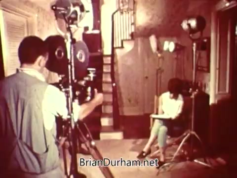 The still depicts a filming set for a National Safety Council public service announcement (PSA). A cinematographer is positioned with a 16mm camera aimed at a seated individual in a casual setting, possibly a living room. The backdrop features stairs and a home-like environment with studio lights illuminating the scene. The atmosphere suggests a focused recording session aimed at conveying a safety message.