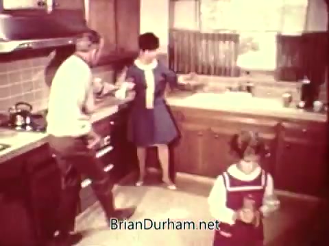 The still features a kitchen scene with a man and a woman engaged in a conversation, while a young girl stands nearby. The aesthetic is typical of mid-20th century domestic settings, highlighting familial interactions. The overall tone suggests a focus on safety and awareness within the home environment, as is common in public service announcements.