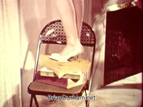 The still shows a foot, wearing a white shoe, positioned precariously atop a stack of books placed on a chair. The setting suggests a domestic environment, possibly illustrating unsafe behavior. The image is part of a public service announcement by the National Safety Council, likely aimed at promoting safety awareness in everyday situations.