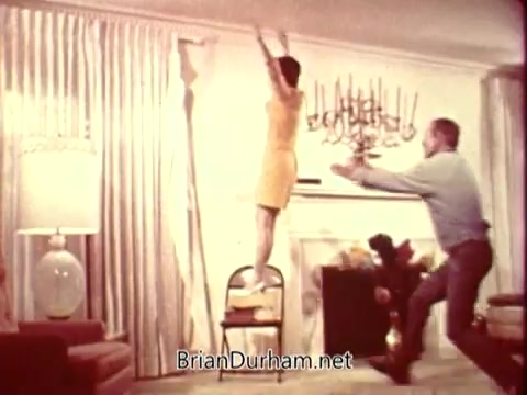 The still from the 'National Safety Council PSA' features a woman in a yellow dress precariously perched on a chair, appearing to reach for something above her. A man is shown leaping towards her, seemingly to catch or support her, highlighting a moment of tension and concern for safety. The setting is a domestic living room, indicated by the decor and furniture.