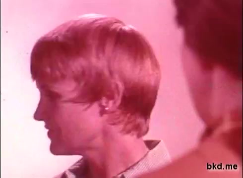 The still features a man with light, medium-length hair styled in a 1970s fashion, shown in profile. The background has a soft pink hue, typical of the era's aesthetic. Another figure is partially visible, possibly engaging with the man. The overall tone is nostalgic and colorful, reflecting the style of commercials from that decade.