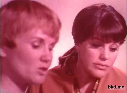The still features two women engaged in conversation, likely discussing the benefits of the Biz detergent. The image is characterized by a soft, warm color palette typical of 1970s advertising, with a slight grain indicative of 16mm film. Their expressions reflect interest or contemplation, suggesting a promotional tone aimed at highlighting product effectiveness.