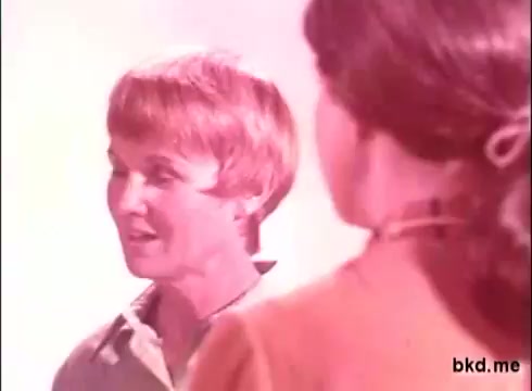The still features two women engaged in conversation, set against a soft, pastel background typical of 1970s advertising aesthetics. One woman has short, blonde hair and is wearing a light-colored blouse, while the other has longer hair tied back and is dressed in an earthy-toned outfit. The scene radiates a warm and inviting atmosphere, characteristic of commercial visuals from that era.