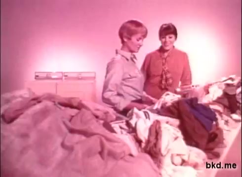 The still features two women engaging with a large pile of laundry, likely discussing the benefits of Biz detergent. The background has a soft pink hue, typical of 1970s advertising aesthetics. The women appear to be friendly and approachable, dressed in period-appropriate clothing. One woman is holding a garment, and both seem engaged in conversation, emphasizing the domestic and practical theme of the commercial.