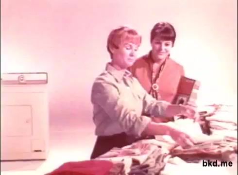 The still from the 'Biz detergent commercial - 1970s' features two women engaged in a discussion over freshly folded laundry. One woman, with short blonde hair, is demonstrating the product, holding a box of Biz detergent, while the other woman looks on attentively, wearing a colorful outfit. The setting has a simple, light background, emphasizing the detergent and the laundry, typical of 1970s commercial aesthetics.