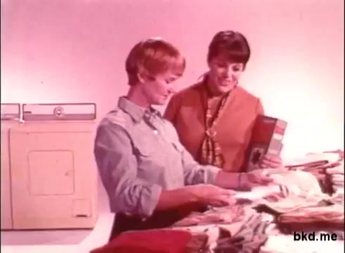 The still from the 1970s Biz detergent commercial features two women in a laundry setting. One woman, wearing a light blue shirt, examines a pile of colorful clothing, while the other, dressed in an orange-brown outfit, smiles and holds a box of Biz detergent. The background is a soft pink, and a washing machine is visible to the left, creating a nostalgic, domestic atmosphere typical of the era's advertising.