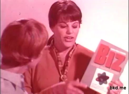 The still showcases two women in a brightly colored setting, discussing a product called 'Biz.' One woman holds a box with the product name prominently displayed, while the other looks on attentively. The overall aesthetic reflects a 1970s style, characterized by vibrant colors and retro fashion. The focus is on the product and its appeal to consumers.