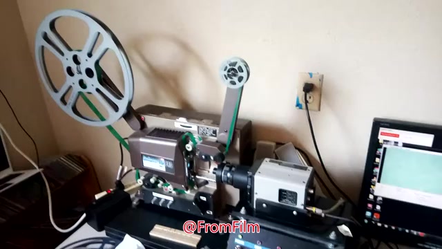 The still shows a setup for transferring 16mm film to video. It features a film projector with large reels, connected to a capture device, and a computer displaying the video output. The arrangement highlights the technical process of digitizing film, emphasizing the equipment used.