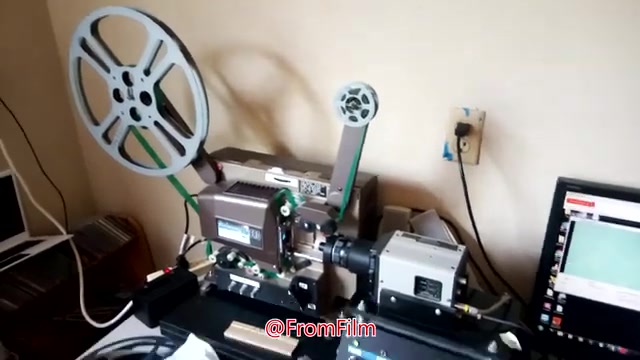 The still shows a 16mm film transfer setup. In the foreground, a film projector is positioned with a reel on the left and electronic components attached to it. There's an interface for capturing the video output on a nearby computer, indicating the film-to-video transfer process. The environment appears casual, suggesting a home studio setup.