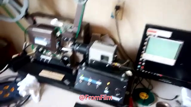 The still depicts a film transfer setup for converting 16mm film to video. Key elements include a film projector and scanning equipment on a workbench, with control panels and a computer monitor displaying live feed or settings. There are also tools and materials like gloves and film reels, indicating an active workspace. The environment suggests a focus on precision in the film transfer process.