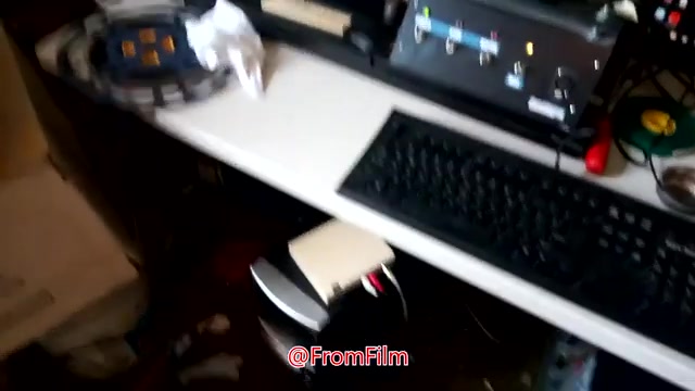 The still depicts a cluttered workspace focused on the film-to-video transfer process. In the foreground, a computer keyboard and a compact device are visible, possibly part of the transfer equipment. A reel of 16mm film is positioned nearby, indicating the medium being processed. The setup suggests a hands-on, technical environment dedicated to the art of film preservation and conversion.