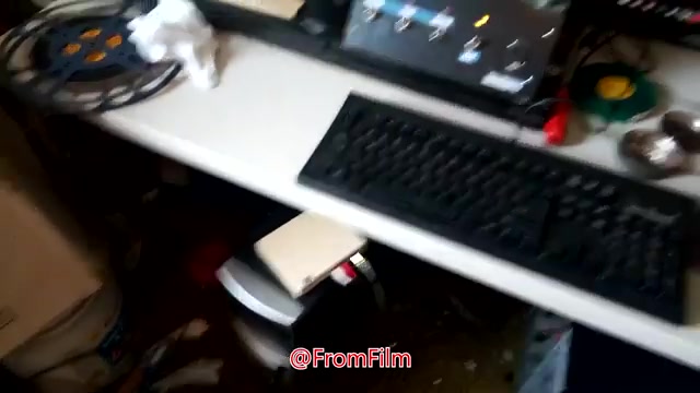 The still captures a cluttered workspace, showcasing equipment used for transferring 16mm film to video. A film reel is prominently displayed, alongside a keyboard and various tools scattered across the desk. The background features monitoring devices, suggesting an active film transfer process.
