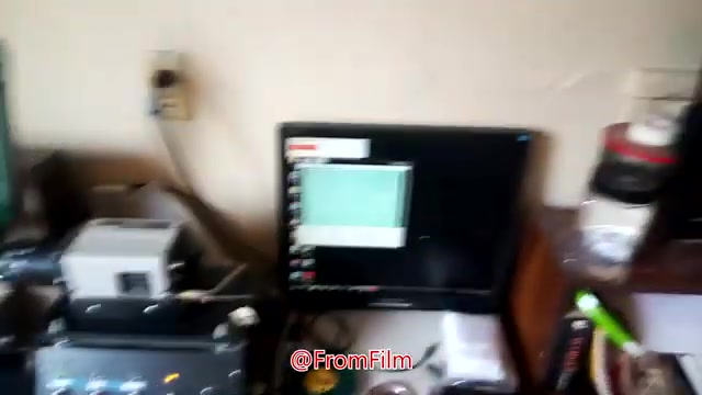 The still captures a cluttered workspace for film-to-video transfer. A film projector is positioned beside a computer setup, displaying a digitally processed image. The scene suggests a hands-on environment, emphasizing the technical aspects of transferring 16mm film. There are also various tools and materials visible, indicating an active film conversion process.