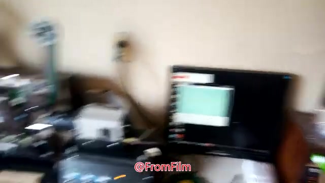 The still captures a cluttered workspace where the film-to-video transfer process takes place. Visible equipment includes a film projector and a computer displaying a software interface. Cables and tools are scattered throughout the area, indicating a hands-on, technical environment. The image conveys a sense of activity and focus on the film transfer process.