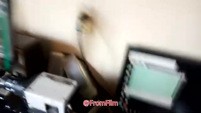 The still shows a workspace focused on film-to-video transfer. In the image, a 16mm film projector is positioned prominently, along with a monitor displaying digital outputs. There are various cables and equipment pieces in the background, reflecting a technical setup for film handling. The environment appears casual and is likely a behind-the-scenes glimpse into the transfer process.