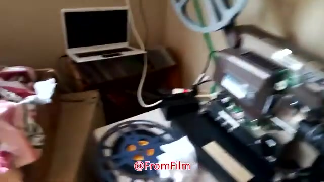 The still captures a behind-the-scenes setup for film-to-video transfer. In the frame, a 16mm film projector is prominently displayed, equipped with reels and an electronic interface. A laptop is positioned nearby, likely used for recording or processing the transferred footage. The workspace appears somewhat cluttered with scattered items, including what seems to be fabric draped over a nearby chair. The environment suggests a practical and functional film transfer operation.