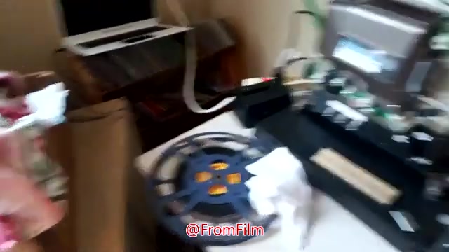 The still shows a workspace for transferring 16mm film to video. In the foreground, there's a reel of film and a transfer machine, likely for digitizing the footage. An open laptop is visible in the background, indicating a digital setup. The environment appears organized yet occupied, suggesting an active project in progress.