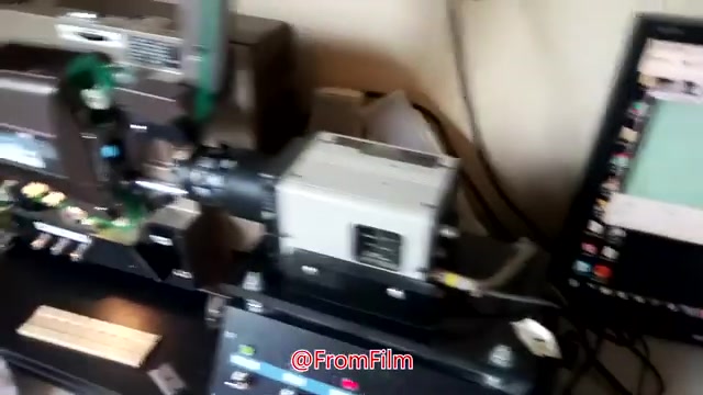 The still features equipment used for transferring 16mm film to video. In the foreground, a film scanner or projector is visible, with connected cables highlighting the setup. A monitor in the background displays the transfer process, suggesting a digital interface for managing the film-to-video conversion. The image overall showcases a behind-the-scenes look at the technical operation involved in the film transfer.