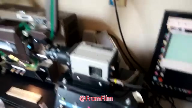 The still shows a behind-the-scenes setup for transferring 16mm film to video. It features specialized equipment, including a film projector and video capture device, connected to a computer displaying digital footage. This highlights the technical process involved in film digitization.