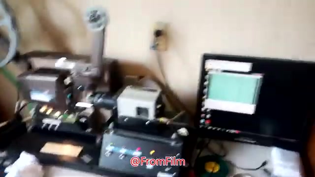 The still features a film transfer setup, showcasing a 16mm film projector alongside a film scanner and a computer monitor displaying the transfer process. The equipment is arranged on a table, with various controls and cables visible, reflecting the behind-the-scenes work involved in transferring film to video. The image captures the technical aspect of film preservation and digitization.